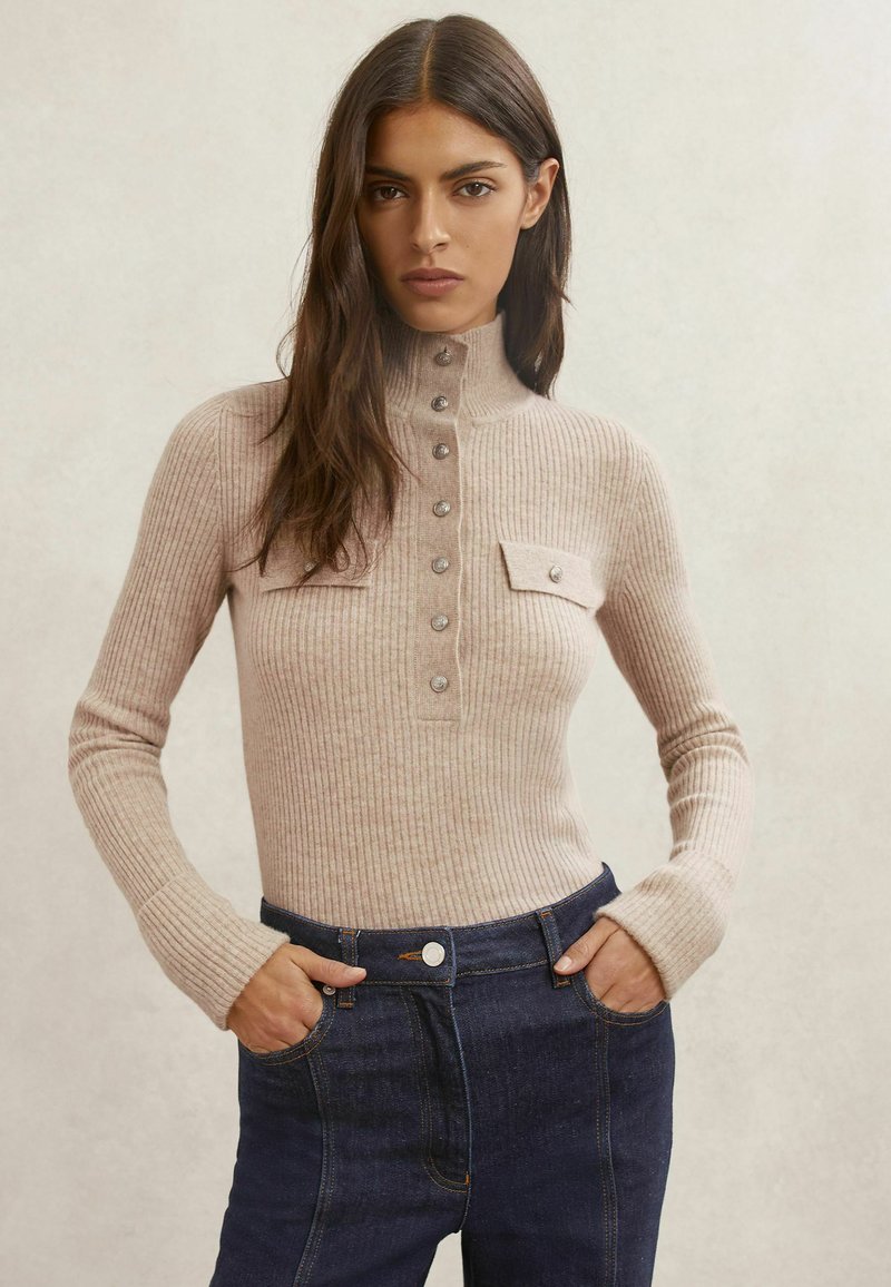 Bézs színű bordázott pulóver magas nyakrésszel, gombos elülső résszel és két elülső zsebbel. Sötét farmernadrággal párosítva, amely testhez simuló derékrésszel rendelkezik.