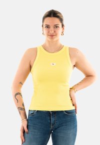 Camiseta de tirantes amarilla con cuello redondo y un pequeño parche de logo, combinada con jeans de mezclilla azules.