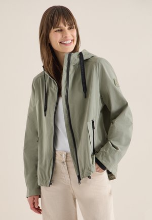 Junge Frau lächelt, trägt eine hellgrüne Kapuzenjacke mit Reißverschluss und schwarzen Kordeln sowie beige Hosen, Hände in den Taschen.