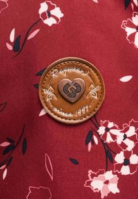 Röd tyg med blommönster; cirkulär brun läderlapp med präglat hjärta och texten "Raquel sedan 1997" med vit sömm detalj.