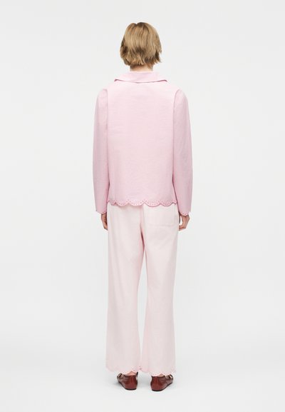 Egy személy áll háttal, világos rózsaszín fodros szélű dzsekit, bőszárú nadrágot és barna cipőt viselve, fehér háttér előtt.