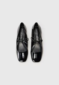 Sapatilhas de balé em couro patentado preto com bico quadrado, apresentando um pequeno laço decorativo no peito do pé. Textura suave e design minimalista.