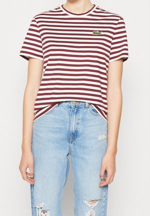 Person iført en rød- og hvidstribet t-shirt med korte ærmer med en lille grøn krokodille-logo og lyseblå revnede jeans.