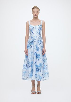 Γυναίκα που στέκεται φορώντας ένα ανοιχτό μπλε με λουλούδια μίντι φόρεμα με φαρδιές τιράντες και ασημένια ψηλοτάκουνα ανοιχτά παπούτσια, πάνω σε λευκό φόντο.