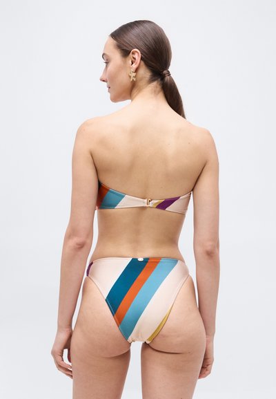 Vrouw met lang haar in een paardenstaart, gekleed in een strapless, meerkleurige bikini met diagonale strepen, van achteren gezien tegen een effen achtergrond.