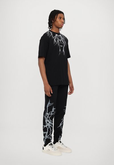 Maglietta nera con grafica e logo a forma di fulmine, abbinata a pantaloni neri con un motivo simile di fulmini. Sneakers bianche completano il look.