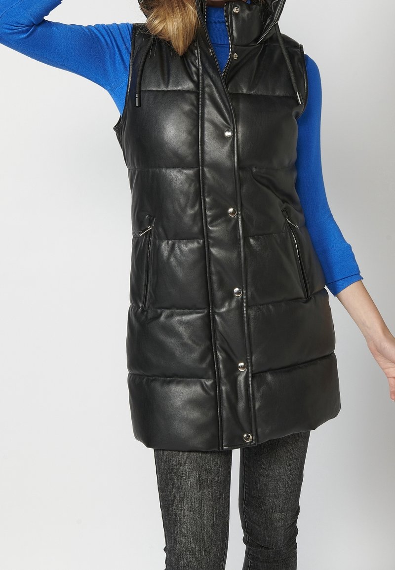 Femme portant un long gilet en cuir matelassé noir avec des boutons-pression, des poches zippées, par-dessus un haut bleu à manches longues et un jean foncé.