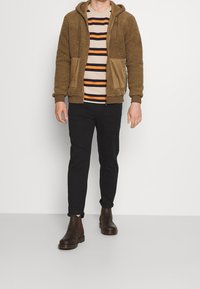 Hoodie in pile marrone con accenti beige, cappuccio e tasche frontali. Indossato sopra una maglietta a righe beige, arancione e blu navy. Pantaloni neri e stivali marroni.