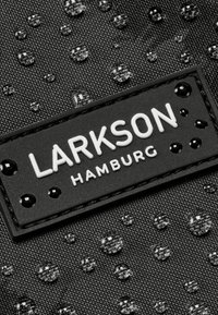 Čierna textúrovaná tkanina s vodnými kvapkami, na ktorej sa nachádza obdĺžniková čierna etiketa s embosovaným bielym textom "LARKSON HAMBURG."