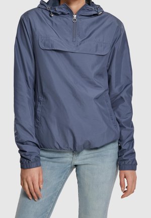 Personne portant une veste coupe-vent à capuche bleue avec fermeture éclair à l'avant et un jean bleu clair sur un fond uni.