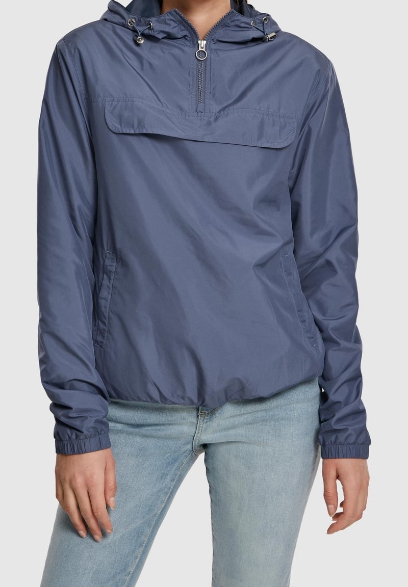 Personne portant une veste coupe-vent à capuche bleue avec fermeture éclair à l'avant et un jean bleu clair sur un fond uni.