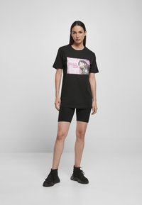 Schwarzes T-Shirt mit einer bunten Grafik eines Charakters und dem Text "MÄDCHEN GEWINNEN." Kombiniert mit schwarzen Radshorts und schwarzen Sneakers.