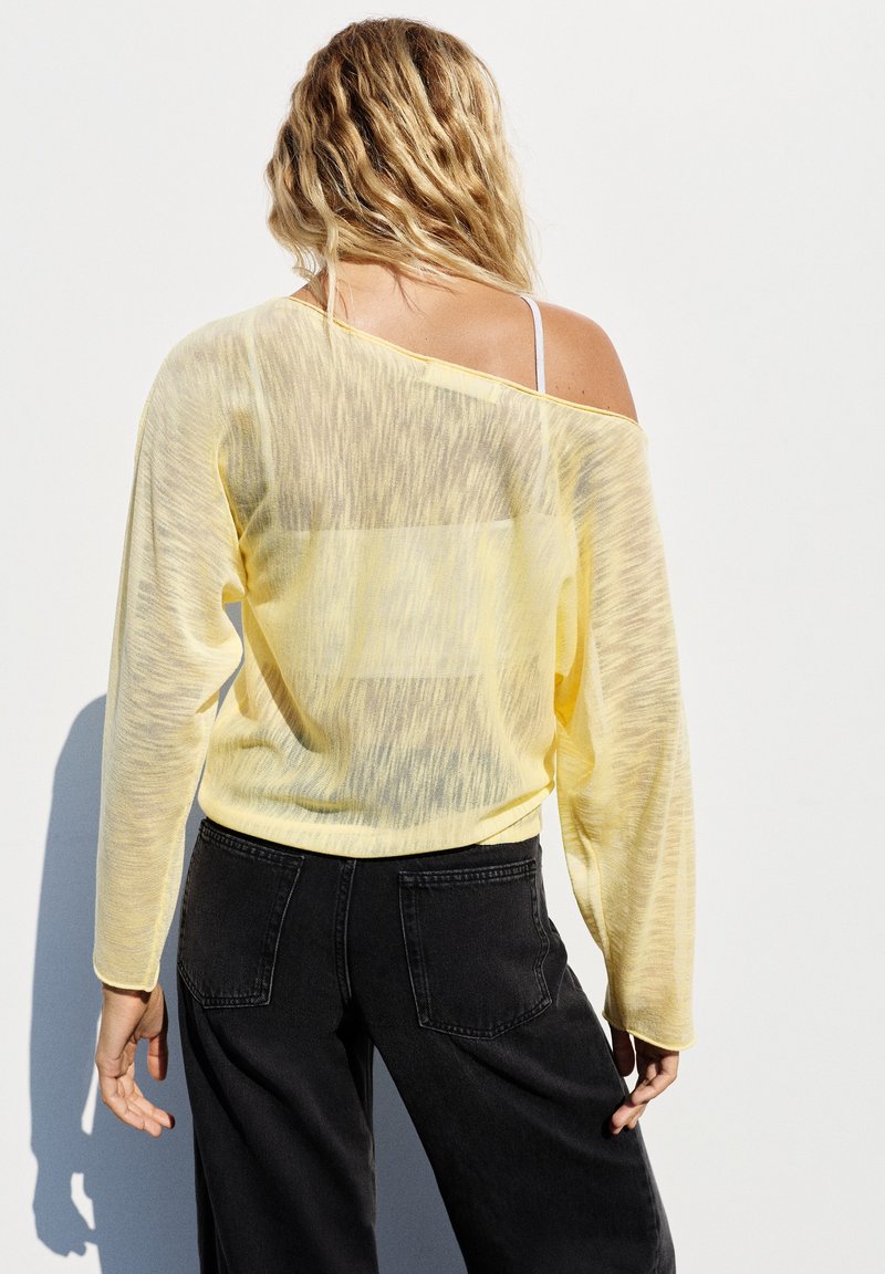 Femme aux cheveux blonds ondulés, vue de dos, portant un haut jaune transparent à épaules dénudées et un jean noir sur un fond clair uni.