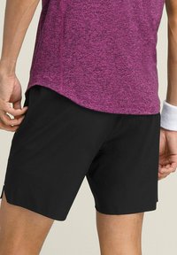 Schwarze Sportshorts mit glatter Textur, seitlichen Schlitzen und lockerer Passform, kombiniert mit einem kurzärmligen, strukturierten lila Oberteil.