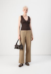 Top aderente marrone con pizzo e nodo frontale, pantaloni larghi color beige, piccola borsa nera e scarpe con stampa leopardata. Sfondo semplice.