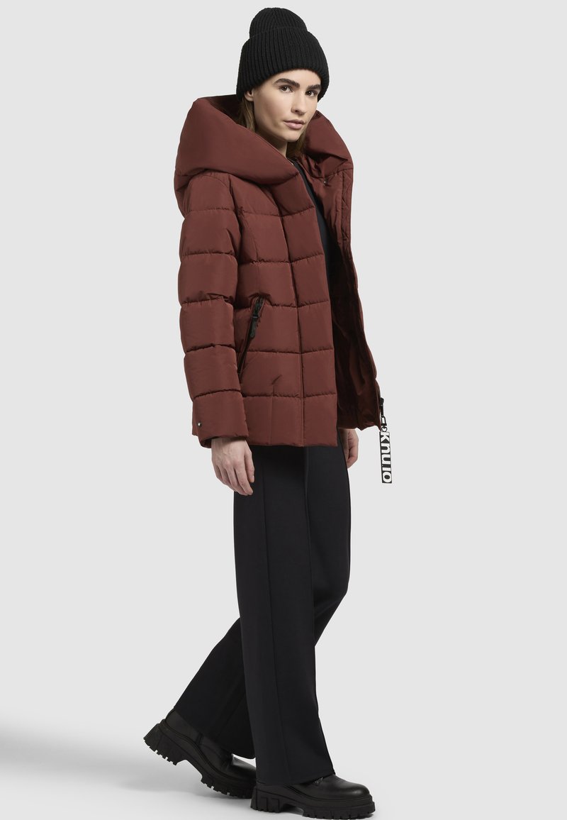 Doudoune de couleur rouille profonde avec une grande capuche, présentant une couture horizontale, deux poches latérales et une fermeture éclair. Associée à un pantalon noir.