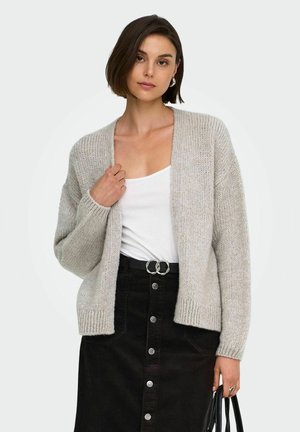 Vrouw met kort bruin haar draagt een lichtgrijze cardigan over een wit topje, zwarte rok met knopen aan de voorkant, houdt een zwarte handtas in haar rechterhand.
