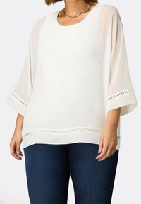 Donna che indossa una blusa bianca con maniche trasparenti a 3/4 e bordi in pizzo, abbinata a jeans blu scuro e una collana in oro.