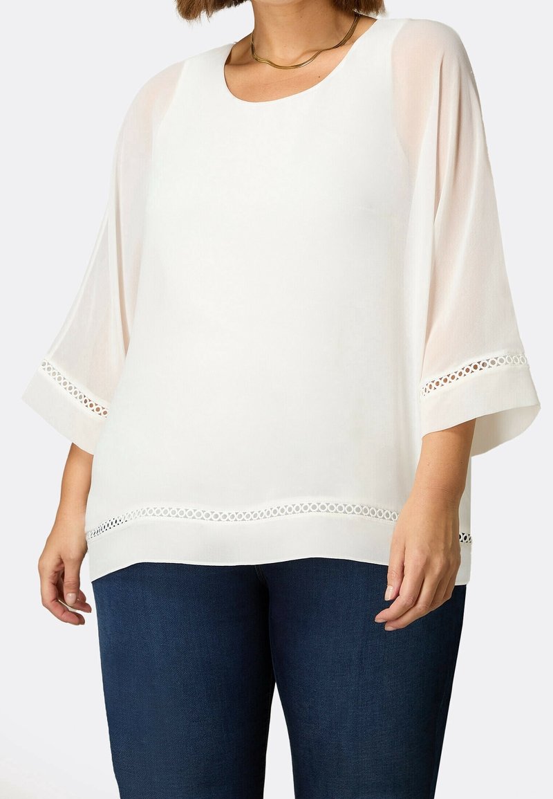 Donna che indossa una blusa bianca con maniche trasparenti a 3/4 e bordi in pizzo, abbinata a jeans blu scuro e una collana in oro.