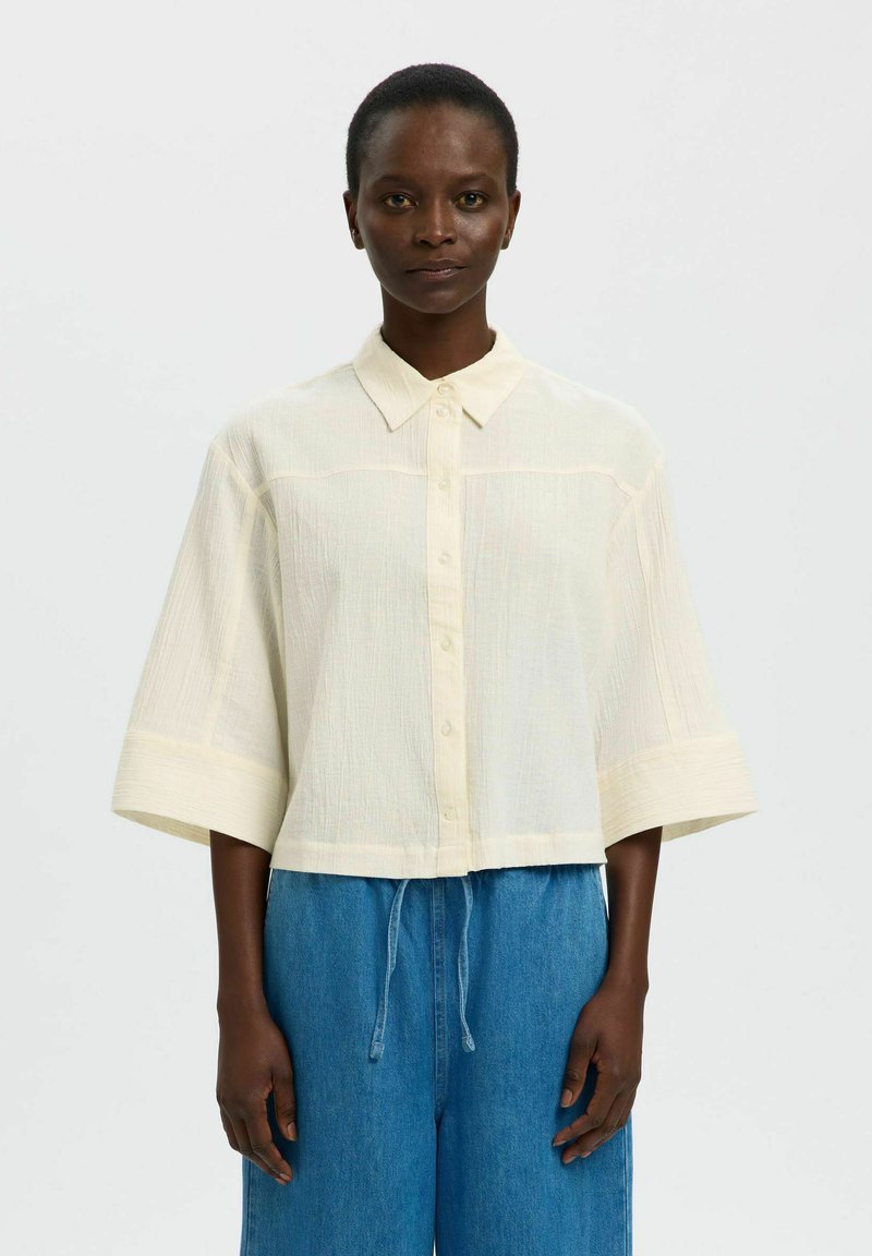 Femme portant une chemise boutonnée ample crème à manches courtes et un pantalon en jean bleu à cordon, debout devant un fond clair uni.