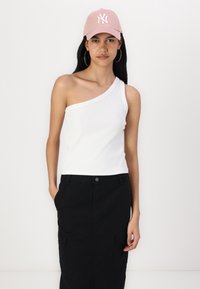 Haut côtelé blanc à une épaule associé à une jupe noire avec des poches, ainsi qu'une casquette rose pâle avec un logo blanc. Le modèle se tient de face.