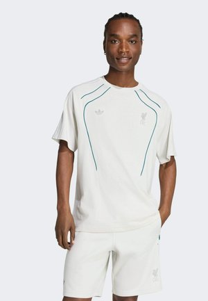 Mężczyzna z warkoczami ubrany w biały sportowy T-shirt Adidas Liverpool FC i pasujące spodenki, stojący z jedną ręką w kieszeni na jednolitym tle.