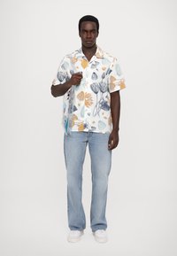 Cămașă cu mâneci scurte, florală, de culoare albă, cu motive florale multicolore. Purtați cu blugi bleu deschis și adidași albi, ținând o geantă.