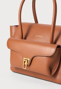 Sac à main en cuir marron texturé avec un rabat, fermoir en métal doré et deux poignées. Présente un logo embossé sur le devant et une forme structurée.