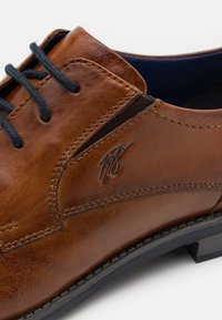 Chaussure en cuir marron avec lacets noirs, coutures discrètes et un logo embossé sur le côté. Texture lisse avec un design à bout arrondi.