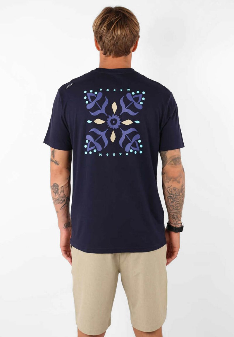 T-shirt en coton bleu marine présentant un motif floral géométrique en violet, beige et aqua au dos. Manches courtes et col rond.