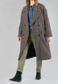 Abrigo marrón de pata de gallo cruzado con solapas anchas y bolsillos laterales, llevado sobre una camisa de denim azul y pantalones verdes, combinado con botas negras hasta el tobillo.