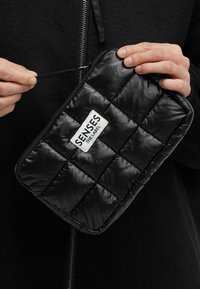 Schwarzes, gesteppte Täschchen aus glänzendem Material mit Reißverschluss und Etikett mit der Aufschrift "SENSES THE LABEL." In der Hand gehalten vor dunklem Hintergrund.