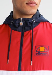 Röd och marinblå vindjacka med dragkedja fram, huva och logodetaljer. Har justerbara dragsnören och blanka detaljer i metall.