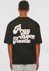 Tricou negru cu mânecă scurtă, având pe spate un text mare alb care spune "Roagă-te, Iubește, Muncă." Confecționat din bumbac, prezentând o croială lejeră.