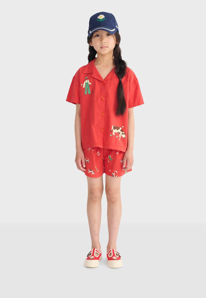 Ragazza con capelli lunghi intrecciati, indossa una maglietta rossa e pantaloncini con stampe di animali da fattoria e contadini, un berretto blu navy con fiore e scarpe rosse.