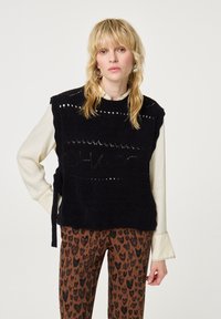 Blonde vrouw die een zwart gebreid vest draagt over een crèmekleurige blouse en bruine broek met luipaardprint, staand tegen een effen lichte achtergrond.