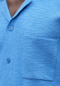 Svetlomodrá košeľa na gombíky s textúrovanou tkaninou, s vreckom na ľavej strane hrudníka a plochým golierom. Gombíky sú rovnakej farby ako košeľa.