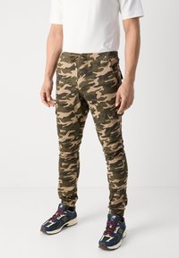 Camouflage cargobroek in groen en beige met elastische boorden, zijzakken en een trekkoord tailleband. Gedragen met multikleuren sportschoenen.