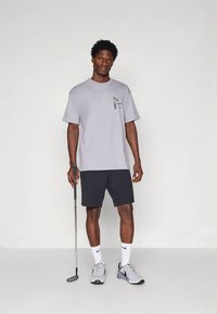 Γκρι βαμβακερό T-shirt με γραφικό σκύλου στο στήθος, σε συνδυασμό με μαύρα σορτς. Το μοντέλο κρατάει ένα γκολφ κλαμπ και φοράει γκρι αθλητικά παπούτσια.