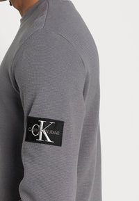 Grå långärmad tröja i texturerad tyg med rund halsringning. Har en svart etikett med "Calvin Klein Jeans" på vänster ärm.