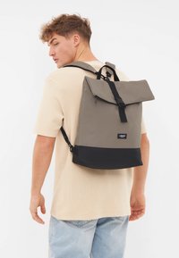 Šedý a čierny látkový batoh s prekrytím na zips, nastaviteľnými popruhmi a štítkom s logom. Materiál vyzerá odolne a je vodeodolný.