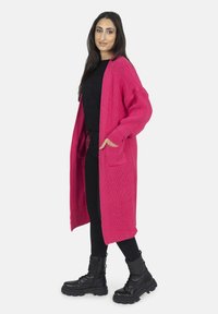 Lång stickad cardigan i klar rosa, med en lös passform, två framfickor och ribbade ärmslut, ihop med svarta kläder och stövlar.