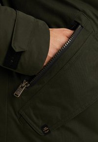 Olivově zelená bunda se zipovou kapsou, vyrobená z texturované látky, s vrchním šitím a malou logovou etiketou. Zip má metalický povrch.