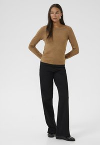 Maglione lungo color beige con scollatura a barca, abbinato a pantaloni neri a zampa d'elefante. Il maglione ha una texture liscia, mentre i pantaloni mostrano una leggera lucentezza.