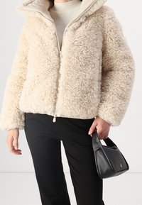 Beige Jacke aus Kunstpelz mit durchgehendem Reißverschluss und Kapuze, weicher Textur und weiten Ärmeln, kombiniert mit schwarzen Hosen und einer kleinen schwarzen Tasche.