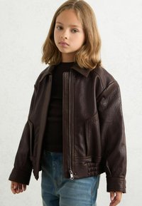 Pruun nahkast jope, millel on tekstureeritud viimistlus, lukuga kinnitamine, terav krae ja elastne äär. Sobitatud musta särgiga ja siniste teksadega.