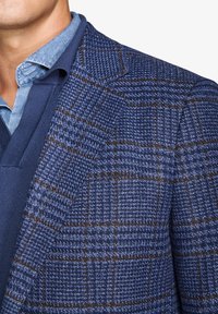 Uld-blandet blazer i blå med et ternet mønster, der har brune accenter. Klippede reverser, tekstureret stof, lagdelt over en blå skjorte.