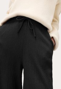 Pantalon large noir à côtes avec une taille à cordon, présentant une surface texturée. Associé à un pull de couleur crème.