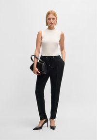 Top bianco a coste senza maniche, pantaloni neri con coulisse e décolleté neri a punta. Il modello tiene in mano una borsa nera strutturata.