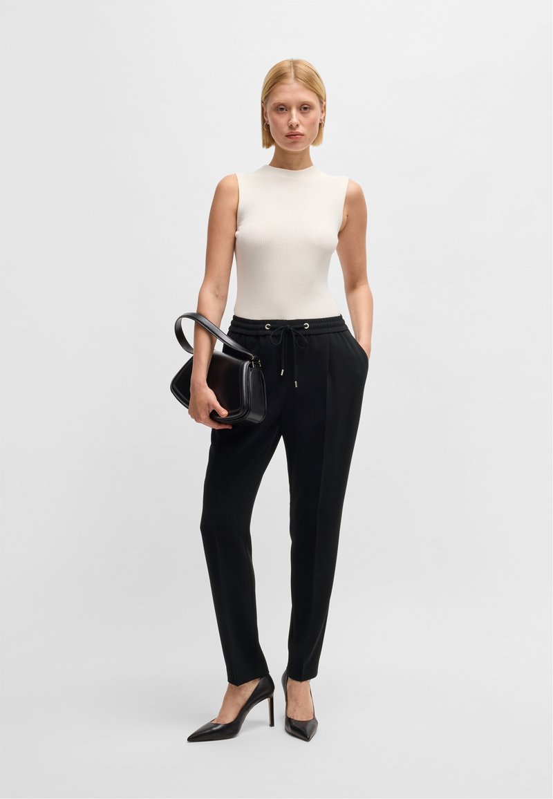 Top bianco a coste senza maniche, pantaloni neri con coulisse e décolleté neri a punta. Il modello tiene in mano una borsa nera strutturata.